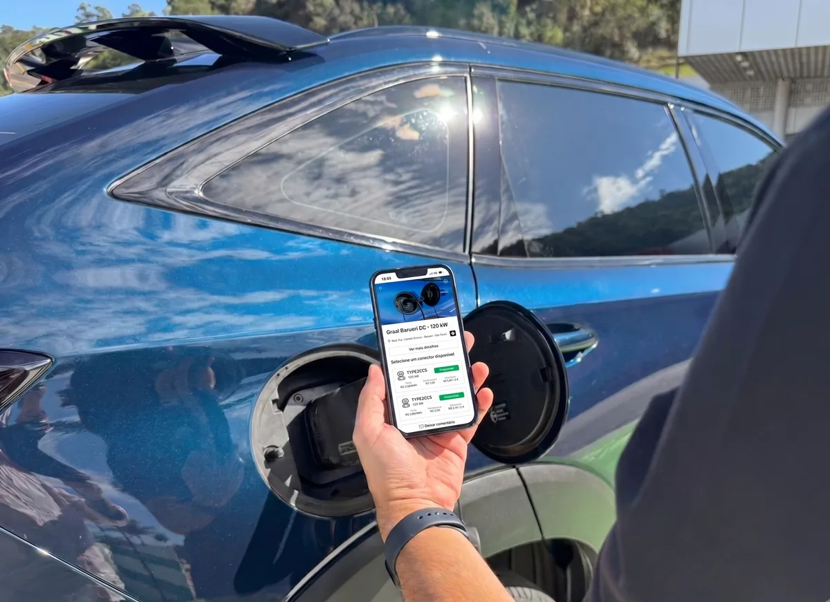 Motorista segura celular com o aplicativo My GWM aberto na tela do carregador Graal Barueri DC de 120 kW, ao lado da entrada de recarga de um SUV azul