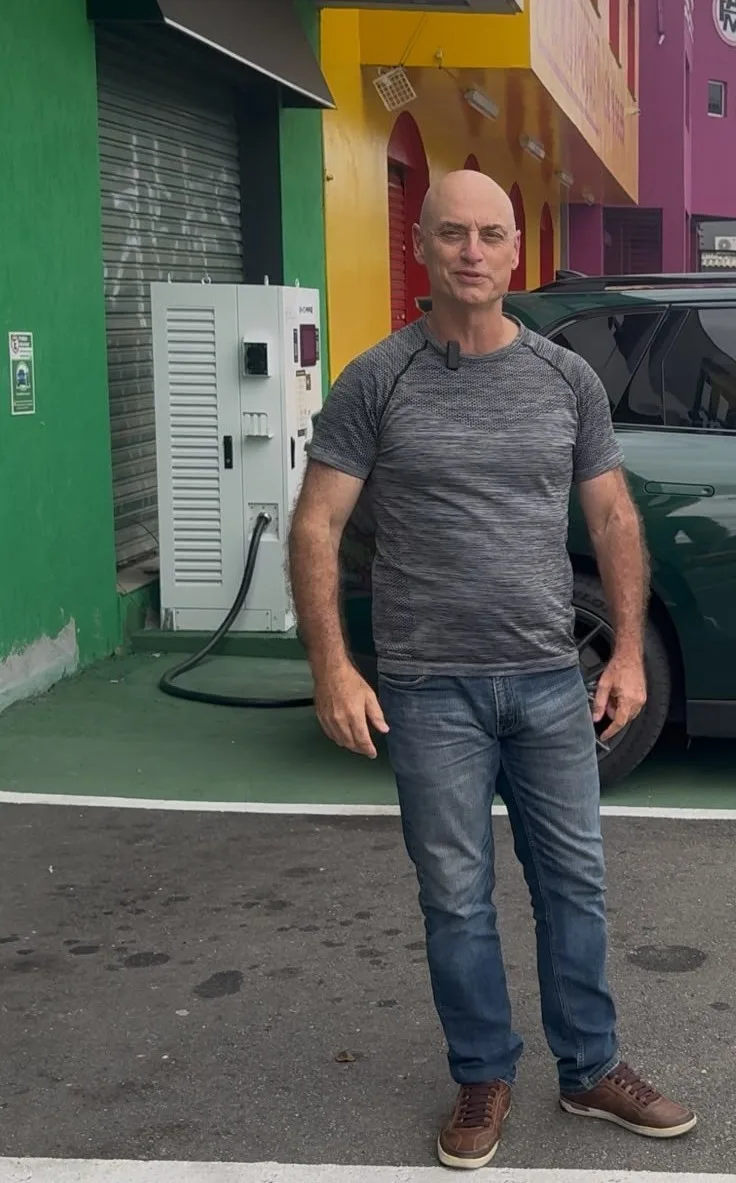 Maurício Barros ao lado de um carregador de parede e de um carro elétrico verde estacionado