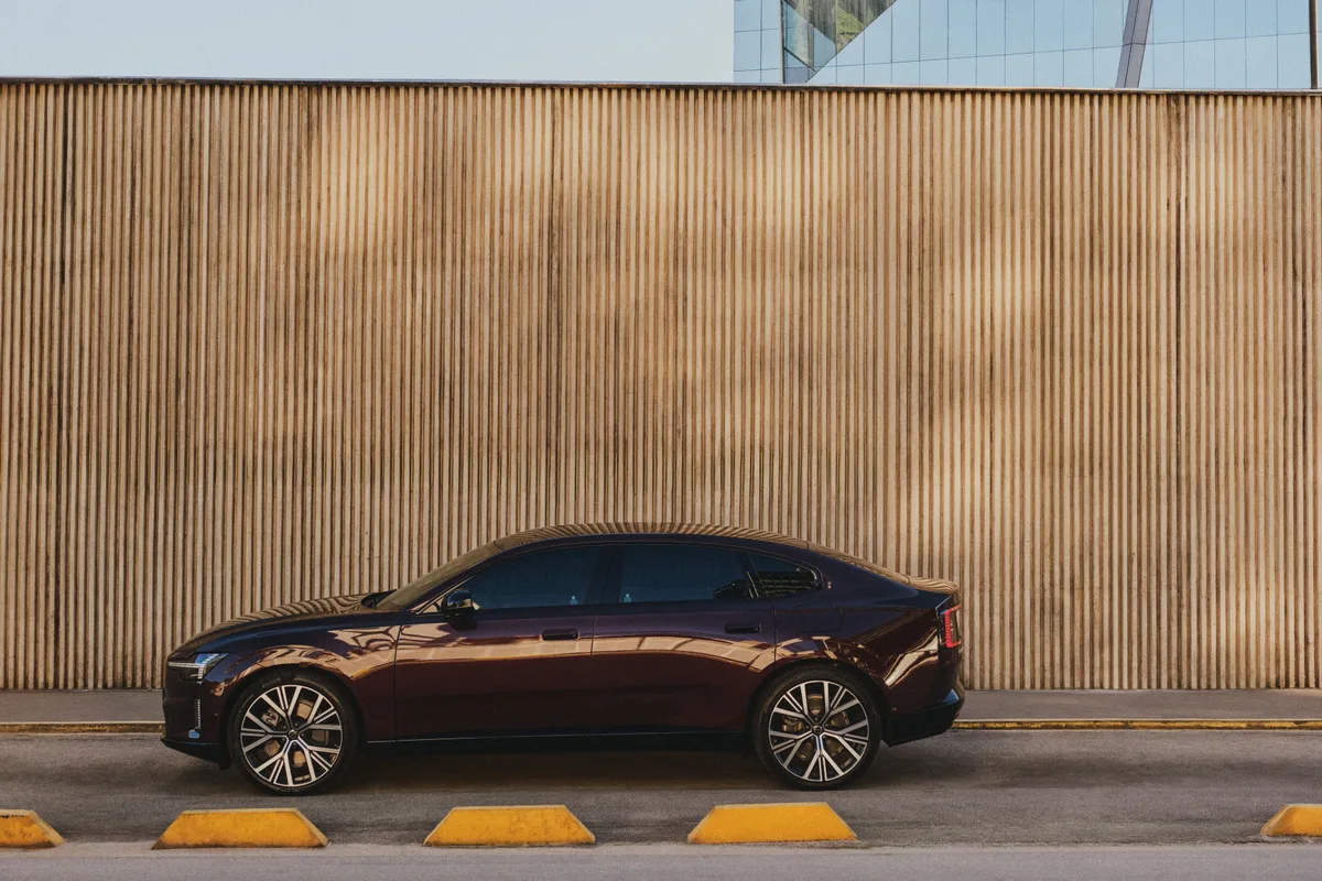 Perfil lateral do Volvo ES90 vinho estacionado diante de uma parede ripada, mostrando a silhueta fastback